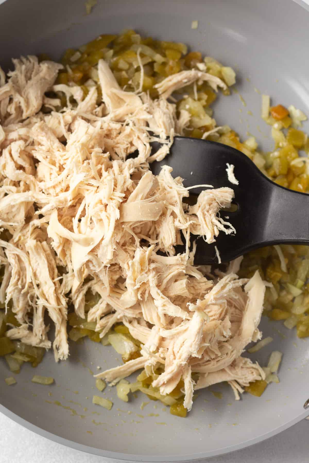 Shredded chicken being stirred into cooked diced onion and green chilis in a gray pan.