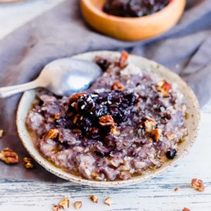 bowl of blueberry crisp oatmeal with a silver spoon in it on the left. a wooden bowl with blueberry jam is in the right background. both bowls are on a gray towel.