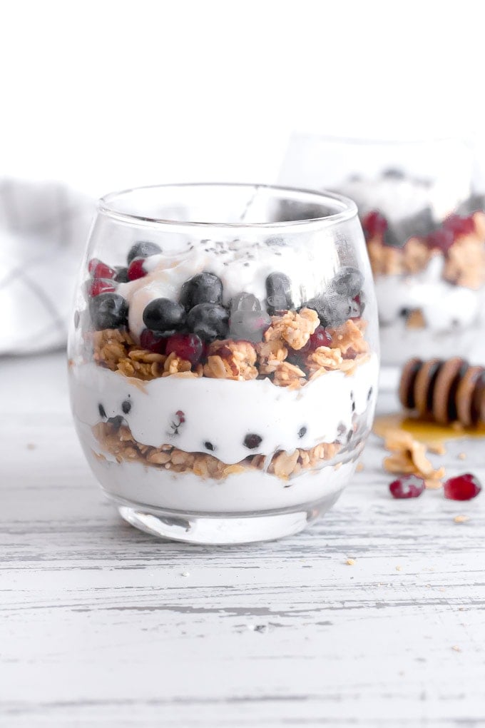 a front shot of a stemless wine glass with breakfast yogurt parfait in it. parfait consists of alternating layers of vanilla yogurt, granola, and blueberries and pomegranate seeds. Another parfait is in the right background and a white towel with thin black outlined squares is the left background. A honey stick lies to the right on the ground, with honey pooling near it.