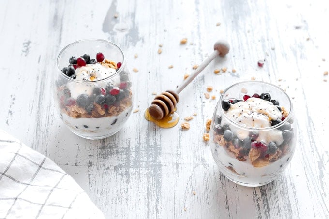 two clear glasses with breakfast yogurt parfait in them,. A white towel with thin black square outlines is in the bottom left corner and a honey dipper with honey drizzling off it is in between the two glasses
