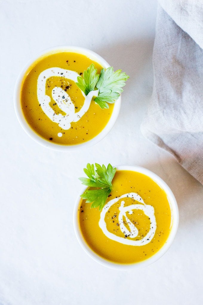 two white bowls with curried butternut squash soup with a swirl of coconut milk and black ground pepper on top. On a slightly gray marble background with a grayish-tan towel to the right.