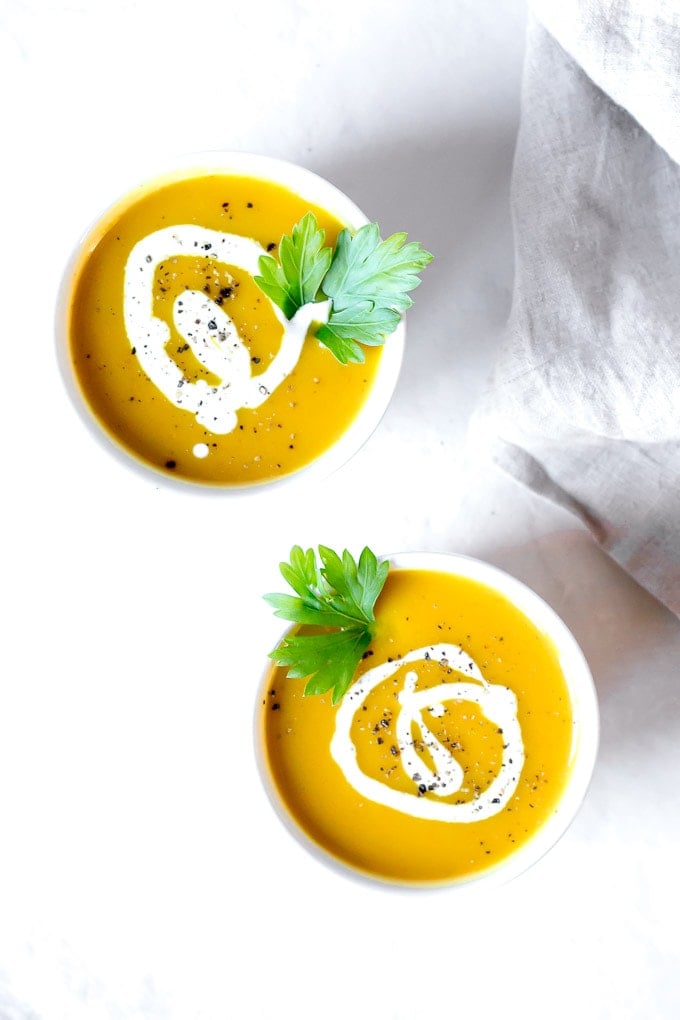 two white bowls with curried butternut squash soup with a swirl of coconut milk and black ground pepper on top. On a slightly gray marble background with a grayish-tan towel to the right.