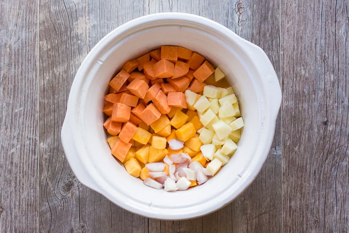top down view of slow cooker insert with cubed butternut squash, sweet potato, apple, and shallot in it, on a gray background