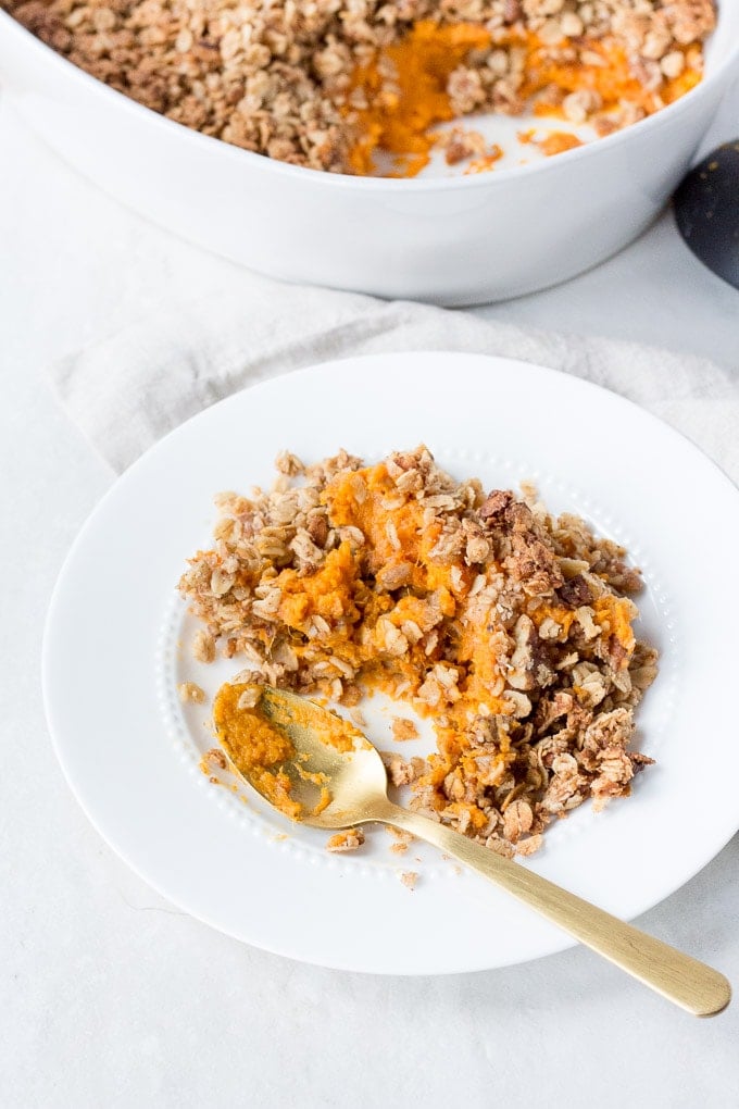 white plate with sweet potato casserole on it and a gold spoon dug into it. A white casserole dish is in the background with a black serving spoon