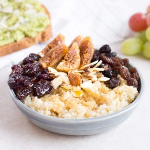 a 45 degree angle shot of quinoa breakfast bowl with avocado toast on a napkin in the upper left, grapes in the upper right, and a napkin and spoon in the bottom right.