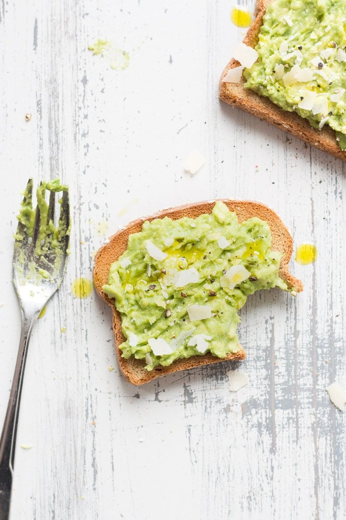 a pieces of gluten free simple avocado toast with a bit out of the bottom right corner. a fork is the left with avocado on the tines and another piece of toast is in the upper right corner. All on a white and gray wood background, from the top down