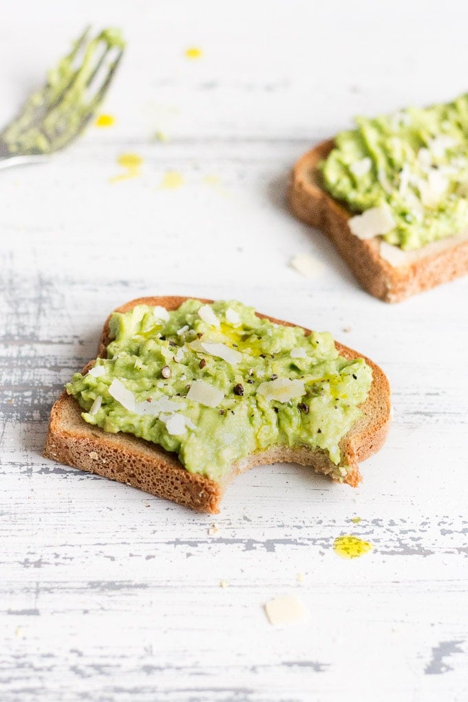 a piece of simple avocado toast with a bit taken out. A fork with avocado on it is in the left background and a second piece of toast is in the right background. The background is a light gray faded wood.