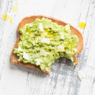 a piece of toast with smashed avocado, salt, pepper, shaved parmesan, and a couple drops of olive oil on a light gray wood background