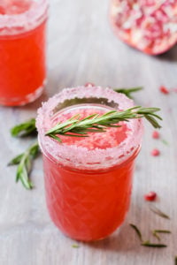 A small mason jar with pomegranate cocktail and a sugared rim. A spring of rosemary lies across the top and there's another cocktail in the left background.