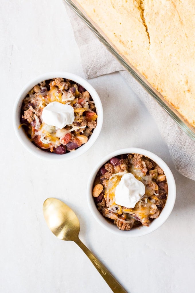 top down view of two white bowls with instant pot beef chili, cheese, and sour cream on top, a gold spoon on the left and a glass dish with cornbread in the upper right