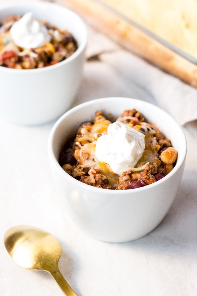 two small white bowl with gluten free beef chili in them, topped with cheese and sour cream next to cornbread and a gold spoon.