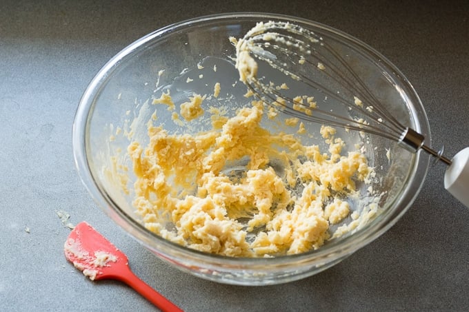 Ingredients for whipped honey butter beaten together in a bowl with a red spatula lying next to it and a hand mixer resting on the lid of the bowl to the right.