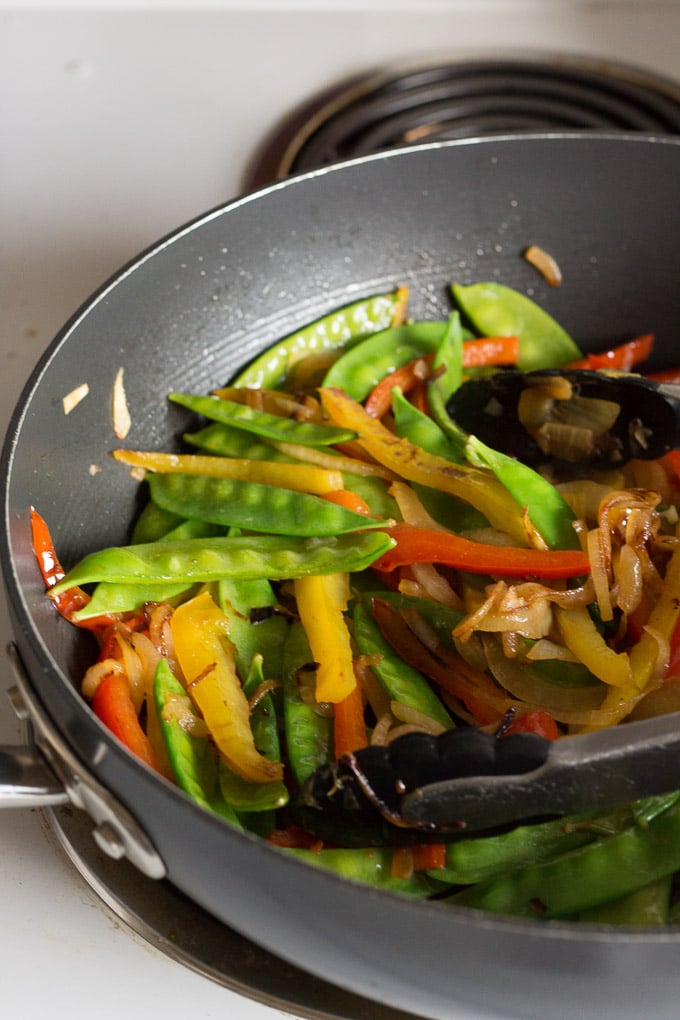 cooked onion and bell pepper slices in a large frying pan with snow peas and a pair of tongs
