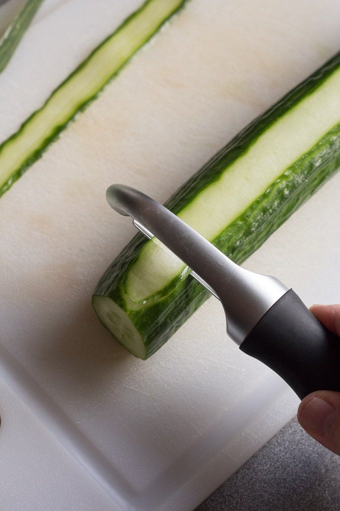 Top down view of a vegetable peeler slicing a cucumber on a white cutting board.