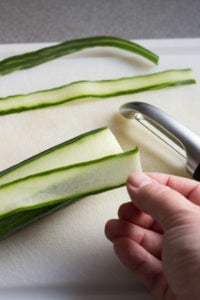 a hand peeling off a cucumber ribbon from the cucumber, with other slices in the back