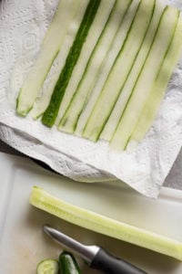 top down view of ribbons of cucumber on a paper towel-lined plate at the top with a peeled cucumber and vegetable peeler on a white cutting board at the bottom