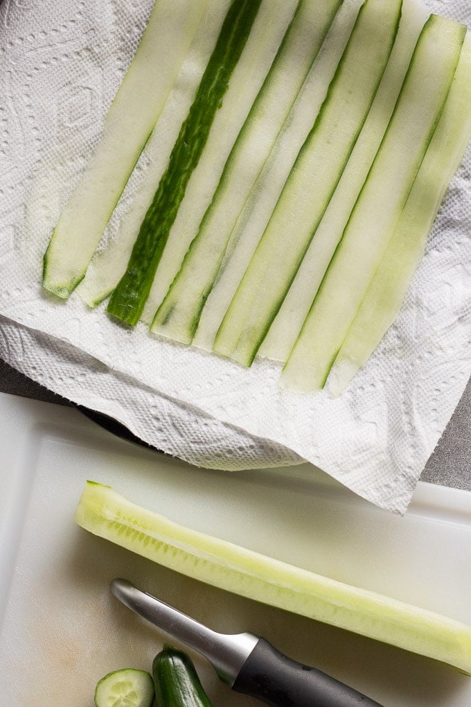 Top down view of ribbons of cucumber on a paper towel-lined plate at the top with a peeled cucumber and vegetable peeler on a white cutting board at the bottom.