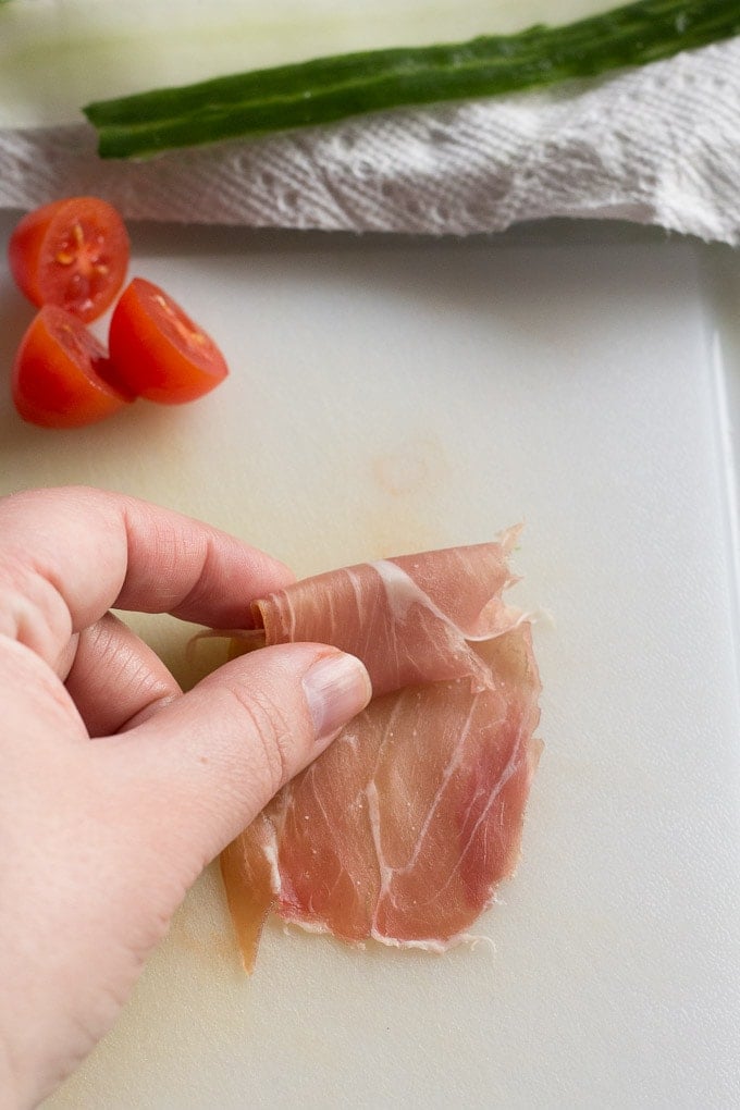 A hand rolling a piece of prosciutto up on a white cutting board with cut cherry tomatoes and cucumber ribbons in the background.