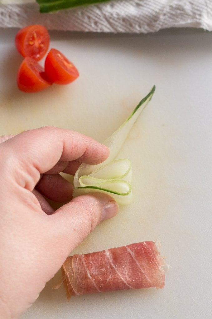 A hand folding a cucumber slice into a ribbon on a white cutting board with prosciutto and cut cherry tomatoes also on the board.