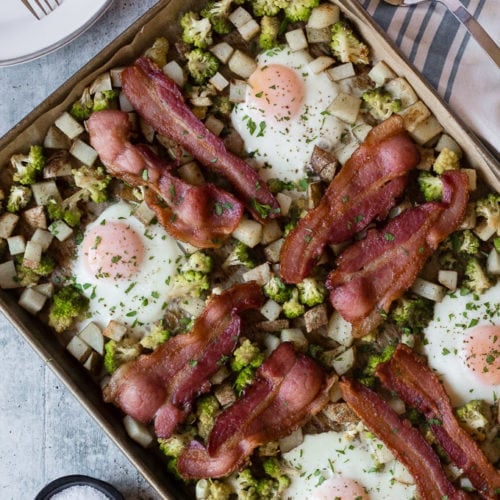 top down view of easy sheet pan breakfast with eggs, bacon, potatoes, and romanesco. Surround by white plates with forks, a striped white and blue towel, and a silver spatula