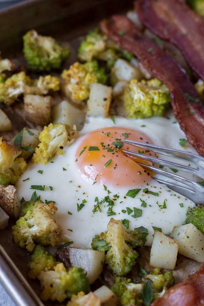 shot of a silver fork cutting into a cooked egg on a sheet pan with potatoes, romanesco, and bacon