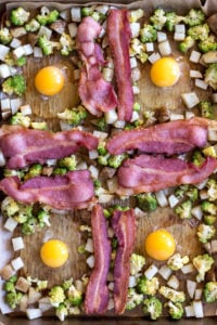 top down view of eggs surround by cubed potatoes, romanesco, and strips of bacon on a sheet pan
