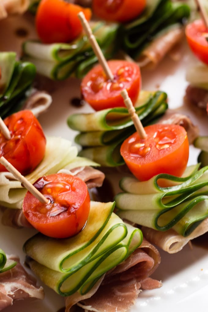 Close up shot of cucumber bites with halved tomatoes, cucumber ribbons, and prosciutto on a toothpick.