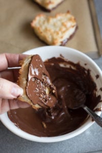 a hand taking out a chewy coconut macaroon heart from a bowl of melted chocolate
