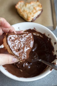 a hand taking out a chewy coconut macaroon heart from a bowl of melted chocolate