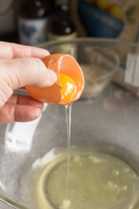 zoomed in shot of an hand holding half an egg shell with the yolk in it and the white dripping out into a large pyrex bowl