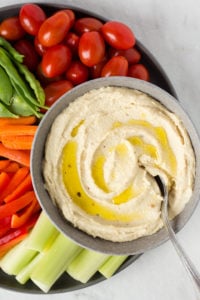 a spoon sits in a bowl of roasted garlic cashew dip, surrounded by veggies on a plate