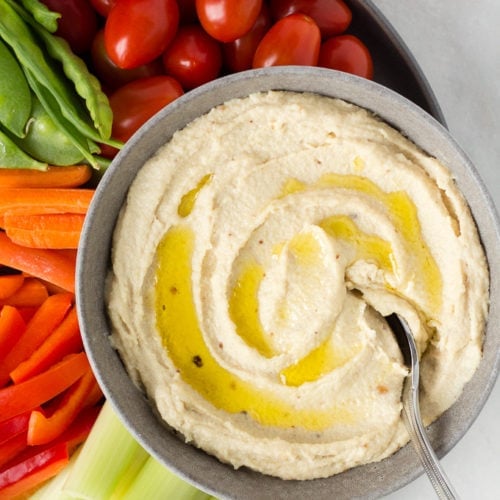 a spoon sits in a bowl of roasted garlic cashew dip, surrounded by veggies on a plate