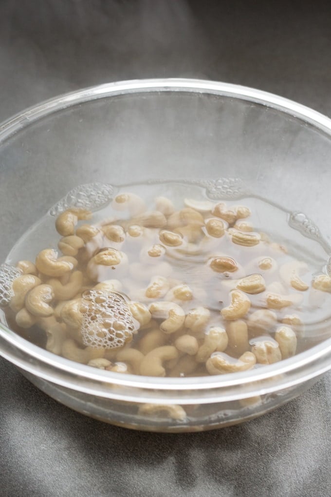 Raw cashews soaking in hot water in a clear bowl.