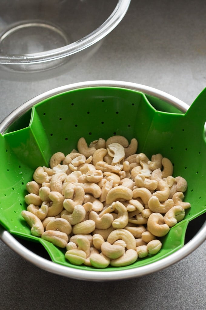Raw soaked cashews in a green colander draining into a silver bowl, all on a gray countertop.
