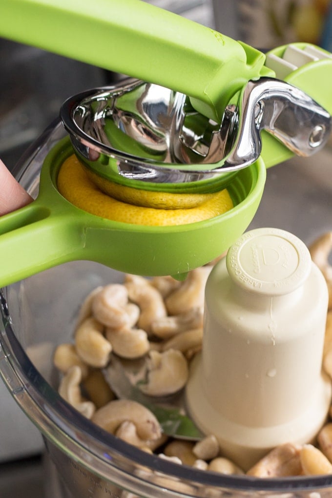 Half a lemon being squeezed by a juicer into a food processor with raw cashews in it.