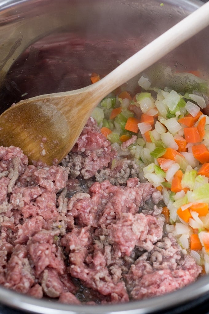 Ground lamb and mirepoix being cooked in an instant pot and stirred with a wooden spoon.