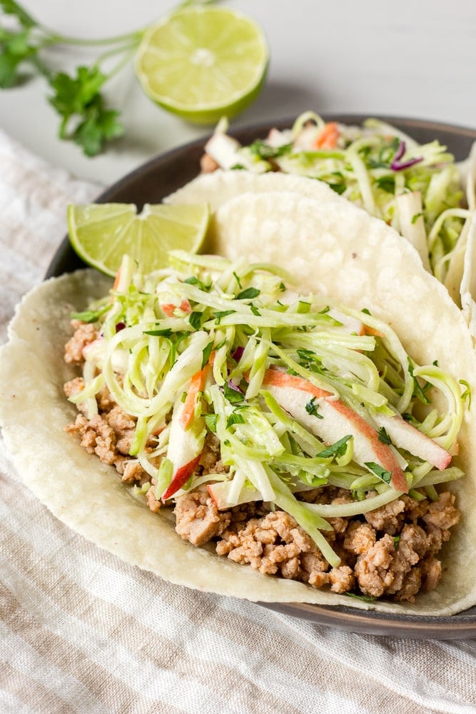 Ground pork tacos with lime on a dark gray plate with a tan and white striped napkin underneath.