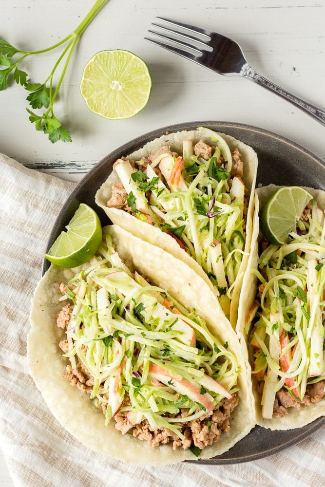 Three soft pork tacos with coleslaw in them on a dark gray plate with sliced lime as garnishes.