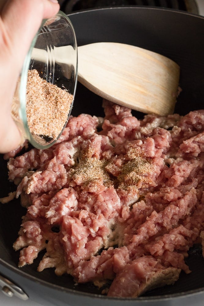 A hand sprinkling seasoning over ground pork in a large black pan.