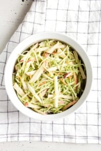 Top down view of apple and broccoli slaw in a white dish with a checked towel underneath.