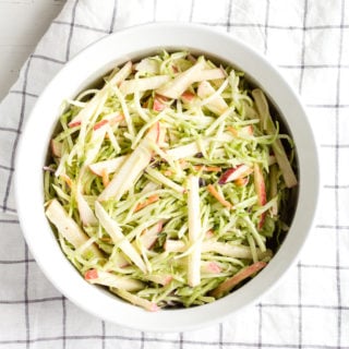 top down view of healthy apple and broccoli slaw in a white dish with a checked towel