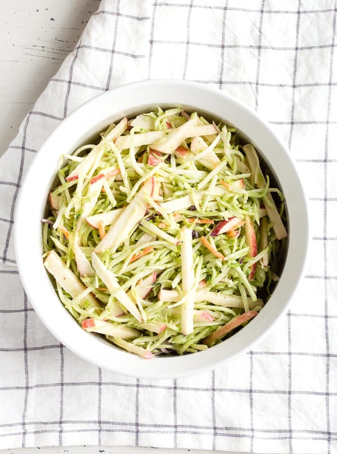 Top down view of apple and broccoli slaw in a white dish with a checked towel underneath.