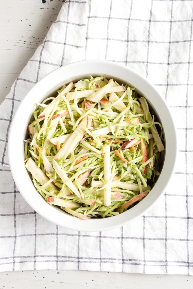top down view of healthy apple and broccoli slaw in a white dish with a checked towel
