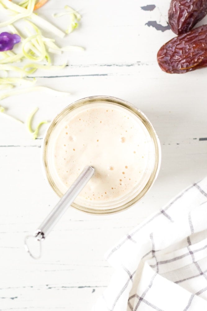 Top down shot of coleslaw dressing in a small mason jar with a whisk coming out, on a white wooden background. A black and white paned napkin is in the bottom right corner.