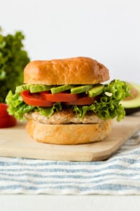 straight on shot of a healthy ground chicken burger with fixing in the background on top of a cutting board