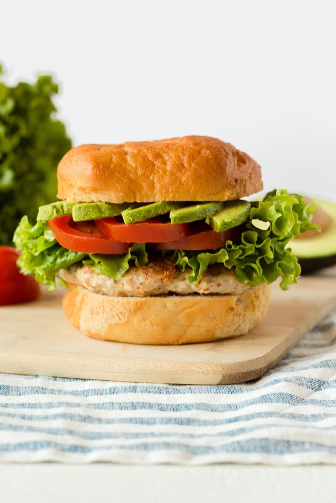 Straight on shot of a healthy ground chicken burger with fixing in the background on top of a wooden cutting board.