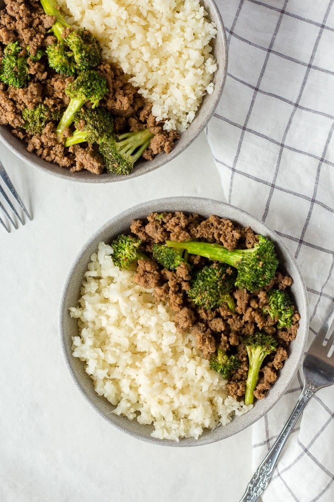 Top down shot of two gray bowls with ground beef, broccoli, and cauliflower rice on a white background.