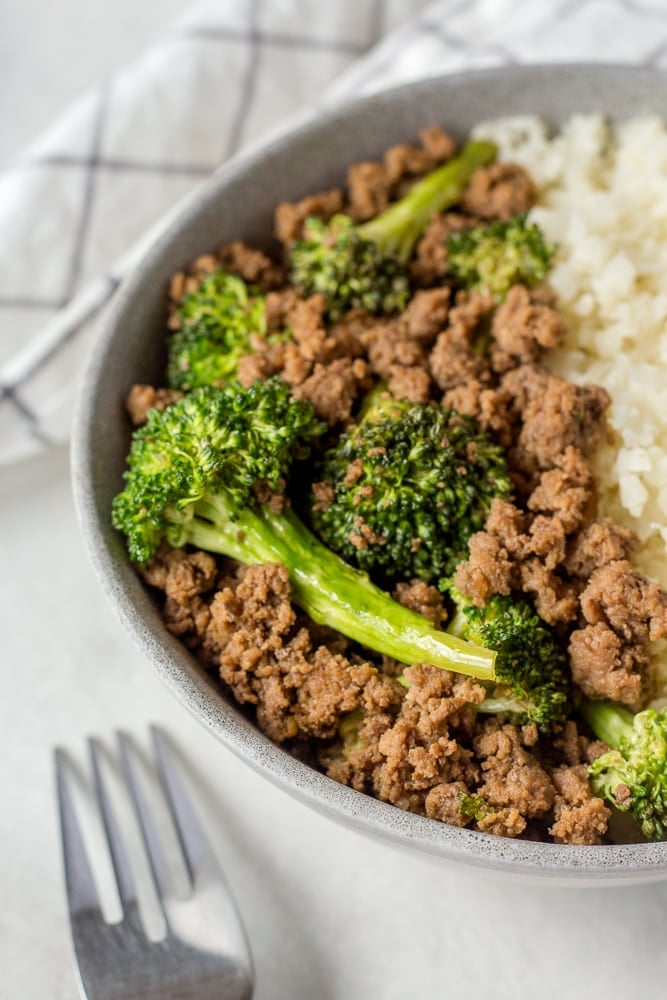 Zoomed in shot of ground beef and broccoli in a gray bowl with cauliflower rice. A silver fork is in the foreground.