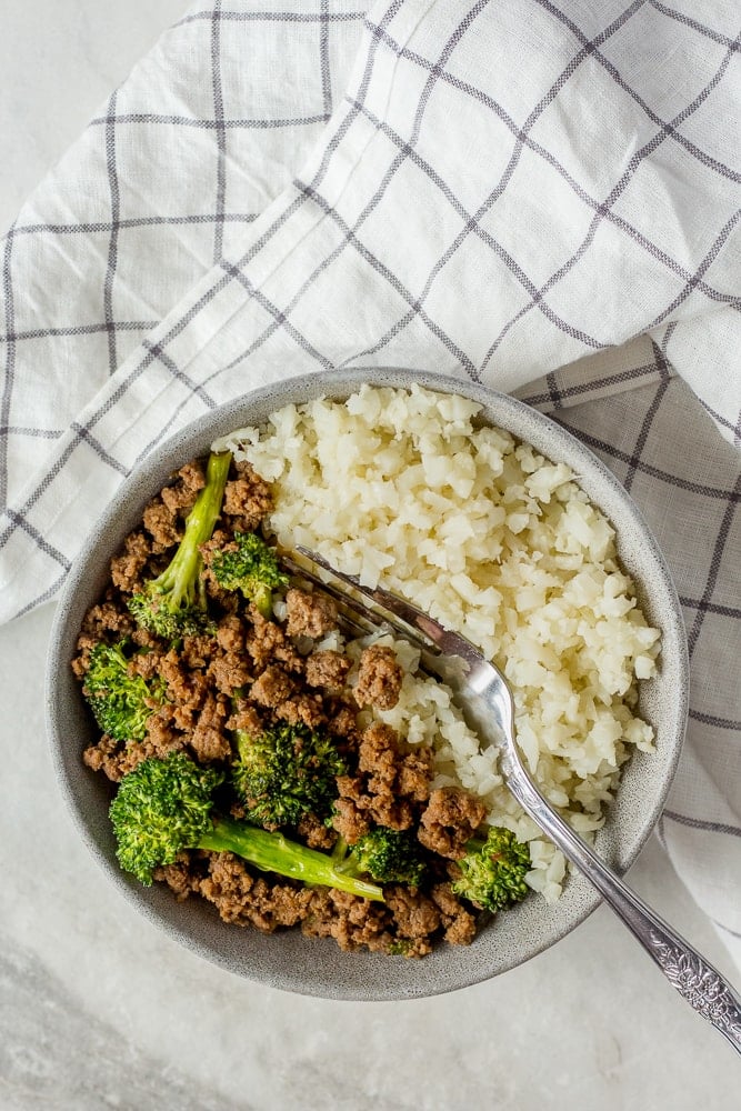 Top down shot of ground beef and broccoli stir fry in gray bowl with cauliflower rice. A fork is in the bowl and a black and white paned cloth napkin is next to the bowl.