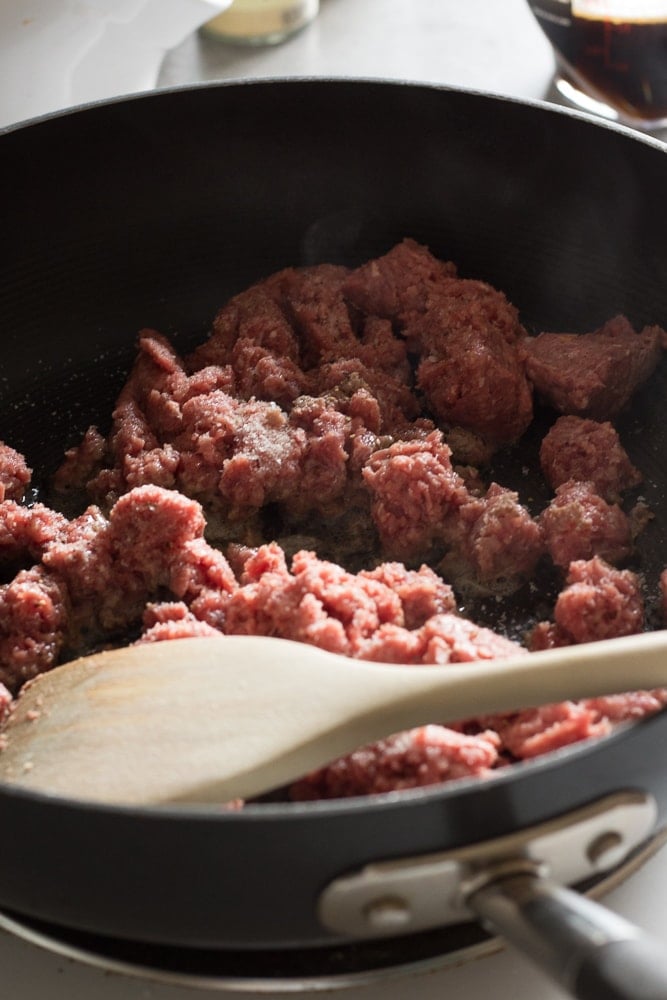 Ground beef cooking on a stovetop in a black pan with a wooden spoon.